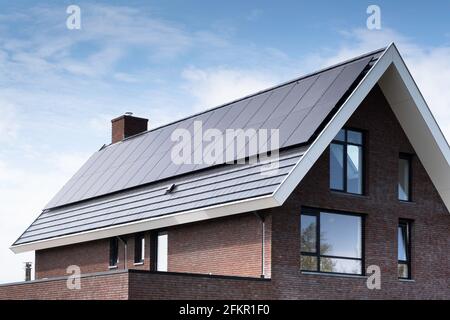 Solar panels mounted on the roof of a modern new-build house in a street in the Netherlands with sun and blue cloudy sky. Sustainable energy Stock Photo