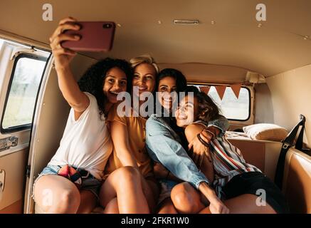 Four beautiful friends taking selfie in a van during a road trip. Smiling women on vacation. Stock Photo