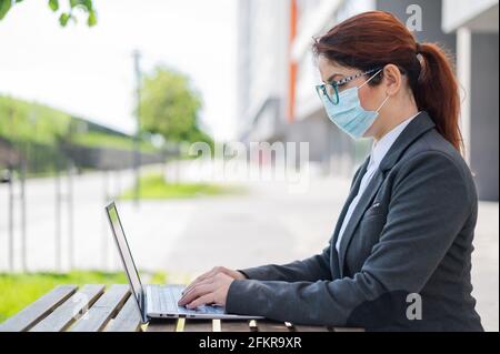 Female manager in a suit works remotely on a computer in a street cafe. Business woman in medical mask is typing on a laptop while sitting on a summer Stock Photo