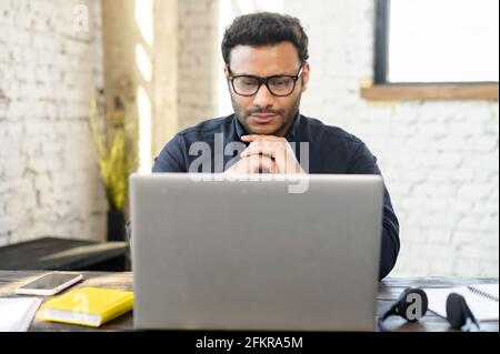 Thoughtful hindu freelancer guy wearing eyeglasses staring at the laptop screen, rested chin with both hands and thinks, concentrated indian man solving difficult tasks, planning business strategy Stock Photo