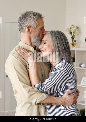 Smiling mid age older couple hugging and kissing standing at home. Stock Photo