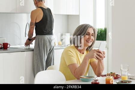 Happy older wife using phone while senior husband washing dishes in kitchen. Stock Photo