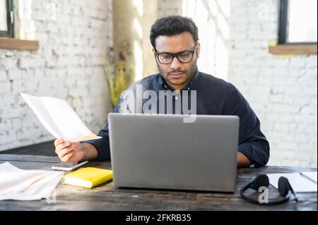 Executive mixed-race male office employee doing paperwork sitting at the desk with laptop in office, focused and serious hindu man in smart casual looking through sheets of documents, preparing report Stock Photo