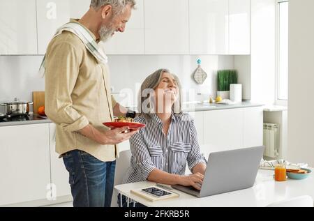 Mature husband bringing breakfast to happy wife working from home office. Stock Photo