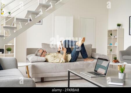Older senior man wearing glasses reading book relaxing lying on couch at home. Stock Photo