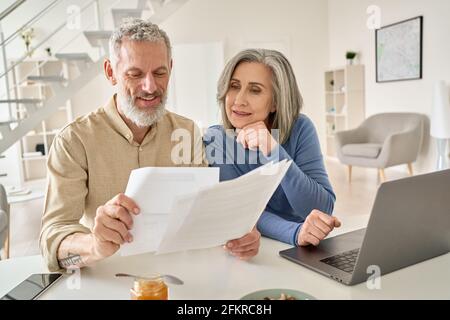 Middle aged couple reading paper bills calculating pension using laptop at home. Stock Photo