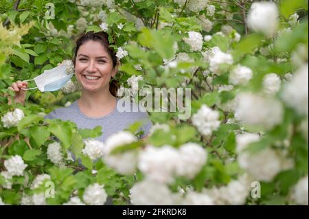 Happy red-haired woman takes off a medical mask while standing next to a blossoming apple tree in a park. Ending seasonal allergic rhinitis. The girl Stock Photo