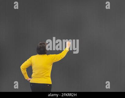 teacher in yellow sweater and black trousers with her back turned writing on a black board with copy space and selective focus Stock Photo