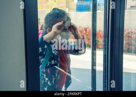 Elementary or kindergarten student wearing a protective face mask looking inward through the window of the school door Stock Photo