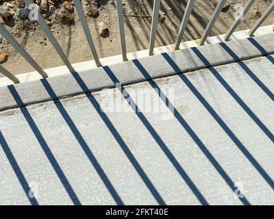 sunlight through a while metal fence with shadows casting on concrete sidewalk diagonal view Stock Photo