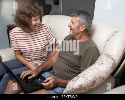 Smiling middle aged couple sitting on their couch using the laptop to buy online at home Stock Photo
