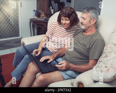 Smiling middle aged couple sitting on their couch using the laptop to buy online at home Stock Photo