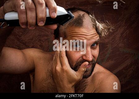 Caucasian man trying to shave his head with an electric razor. A brutal bald man holds a razor in his hand and shaves stubble on a metal background Stock Photo