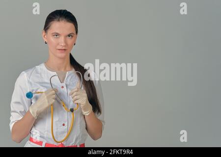 A young female doctor in a white lab coat with a stethoscope in her hands on a grey background Stock Photo