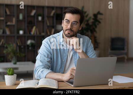 Thoughtful dreamy businessman in glasses sitting at desk with laptop Stock Photo