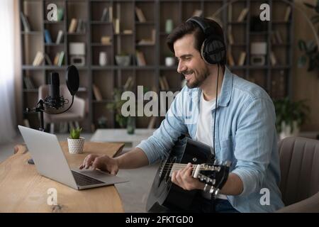 Smiling man in headphones using laptop, playing acoustic guitar Stock Photo