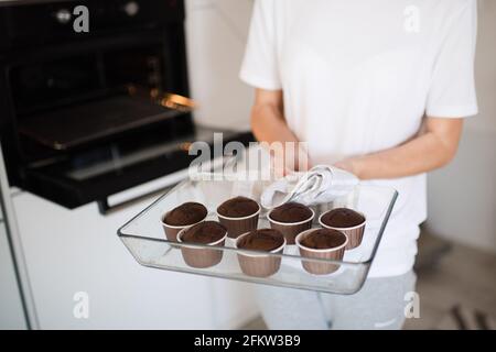 Woman holding glass tray with baked chocolate muffins in kitchen. Fresh out oven. Housewife cook at home. Stock Photo