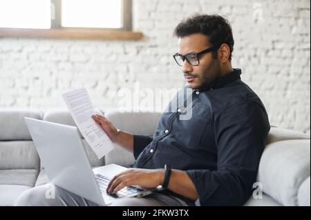Mixed-race indian man in casual shirt sits on the couch, using laptop for remote work, focused freelance guy wearing eyeglasses doing paperwork, analyzing and checking report, preparing business plan Stock Photo