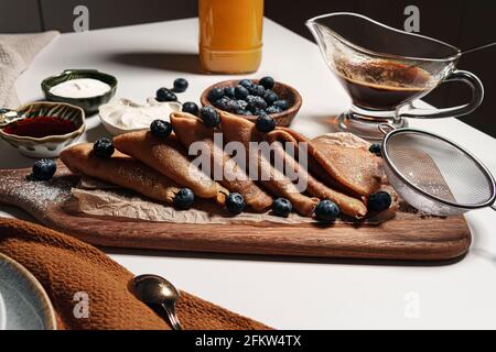 Homemade crepes or pancakes served on the wooden board sprinkled with icing sugar and decorated with blueberry and honey. At the background of a white Stock Photo