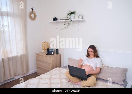 Angry pregnant woman while working on the home bed. Woman showing fist into laptop camera while online remote work Stock Photo
