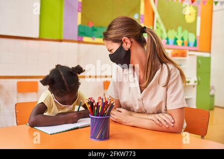 Childminder looks after African girl with her homework with a mask because of Covid-19 Stock Photo