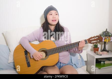 young happy Asian woman playing guitar in bed - attractive and beautiful Korean girl in bedroom enjoying singing and writing music relaxed at home Stock Photo