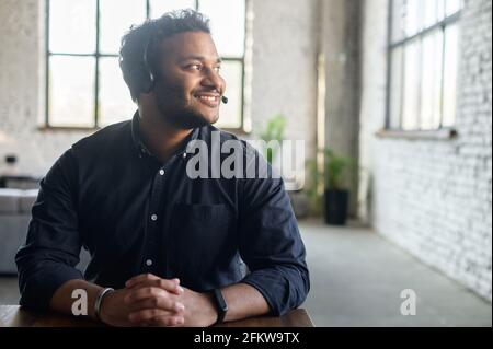 Delighted hindoo man wearing headset device and smart casual shirt looks away dreamily, support representative on the workplace in modern loft office, hindu guy in headphones with mic lost in thoughts Stock Photo