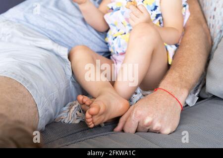 Arm of anonymous dad with protection red wristband on couch with baby daughter. Father sitting on sofa with little girl Stock Photo