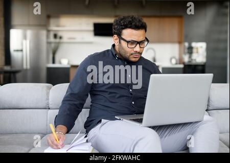 Busy multi-ethnic hindu freelancer guy using laptop computer for work or studying on the distance from home, concentrated indian man watching online webinar and taking notes in notebook, e-learning Stock Photo
