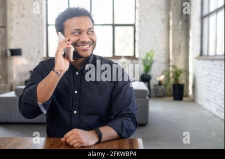 Portrait of cheerful hindu millennial guy chatting on the smartphone sitting at the desk in loft apartment, positive indian man in good mood has phone conversation, talking on the cellphone Stock Photo