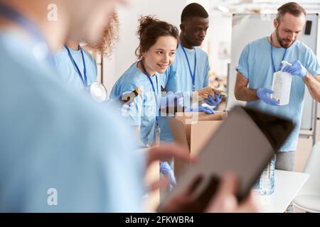 Diverse volunteers in blue uniform sorting foodstuff in cardboard boxes, working together on donation project. Focus on woman Stock Photo