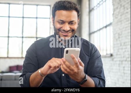 Positive indian guy using smartphone for online communication, smiling mixed-race guy browsing, scrolling news feed, messaging in social media,hindu man holds phone and enjoying new mobile application Stock Photo