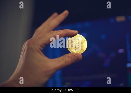 A man holds a gold bitcoin coin in his hand. The graphs of changes in cryptocurrency rates are shown on the laptop monitor Stock Photo