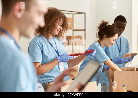 Side view of happy female volunteer smiling, holding pack of pasta while sorting foodstuff in box, working on donation project with team. Focus on Stock Photo