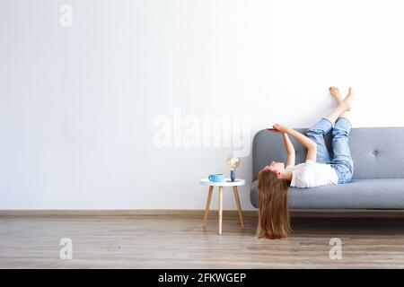 Young beautiful woman wearing white t-shirt on grey textile sofa at home. Attractive slim female in domestic situation, resting on couch in her lofty Stock Photo