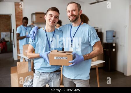 Portrait of two caucasian guys, male volunteers in blue uniform, protective gloves smiling at camera and holding cardboard box for Donation Stock Photo