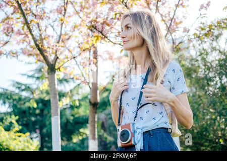 Portrait of a traveling woman with a backpack and a camera in the park with sakura blossoms. A charming young blonde enjoys a walk in a city park on a Stock Photo
