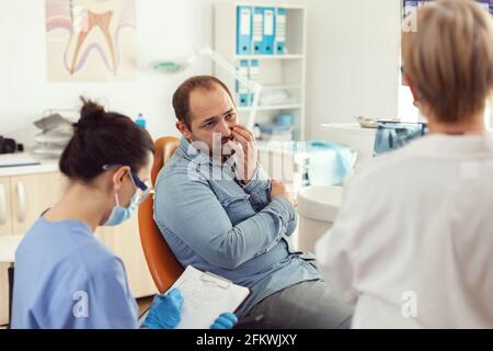 Man patient with tooth pain explaining dental problem to nurse during orthodontic appointment. Assistant writing notes on clipboard while doctor dentist checking stomatology radiography Stock Photo