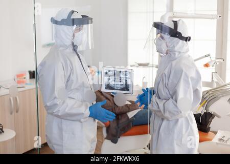 Dentist doctors in overall using tablet explaining dental x ray in stomatologic office during coronavirus. Man wearing face shield and mask showing to nusrse radiography using digital device Stock Photo