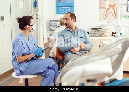 Orthodontist nurse discussing with man patient about tooth consultation writing healthcare treatment on clipboard sitting in medical dental office. Stomatologist making surgery appointment Stock Photo