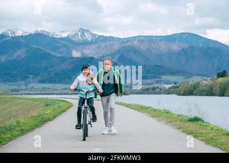 Smiling kids on a bicycle path on the river bank. Brother helping to sister and teaching doing first steps in riding. Happy childhood concept image. Stock Photo