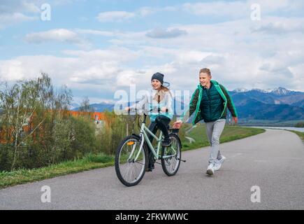 Smiling kids on a bicycle path with snowy mountains background. Brother helping to sister and teaching doing first steps in riding. Happy childhood co Stock Photo
