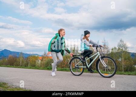 Smiling kids on a bicycle asphalt way. Brother helping to sister and teaching doing first steps in riding. Happy childhood concept image. Stock Photo