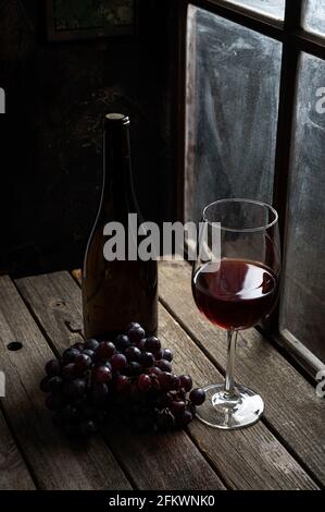 Glass of red wine with grapes and bottle on rustic wooden table next to window Stock Photo