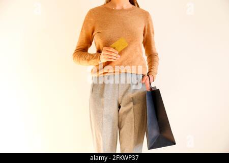 Young slim woman in classic fashionable outfit, brown sweater and loose grey pants went shopping, holding paper bags and vip golden credit / debit car Stock Photo