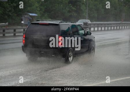 A black SUV is driving in the rain on the highway. The car travels quickly on a wet road. The car drives through a puddle. The car drives forward. It' Stock Photo