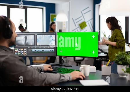 Videographer using computer with chroma key, mock up isolated display editing video and audio footage. Man editor processing film montage on pc with green screen while team working in background Stock Photo