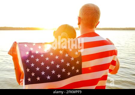 Rear view of young American patriotic couple hugging with USA flag on their backs enjoying beautiful summer sunset by the river. Independence day cele Stock Photo