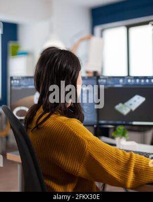 Young woman industrial engineer working at pc with two monitors, screens showing CAD software with 3D prototype of gears metalic mechanical piece. Employee working in creative agency office Stock Photo