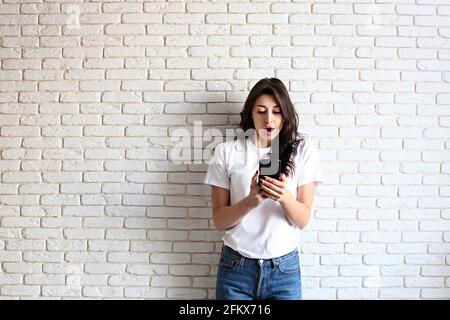 Young attractive woman in blank white shirt received surprise message on cell phone. Pretty female model, looking surprised, open mouth, holding mobil Stock Photo